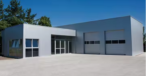 A modern, light-grey prefabricated steel structure, likely a warehouse or commercial building, with large glass entrance doors and two large roll-up garage doors, set on a concrete lot with trees in the background under a clear blue sky.