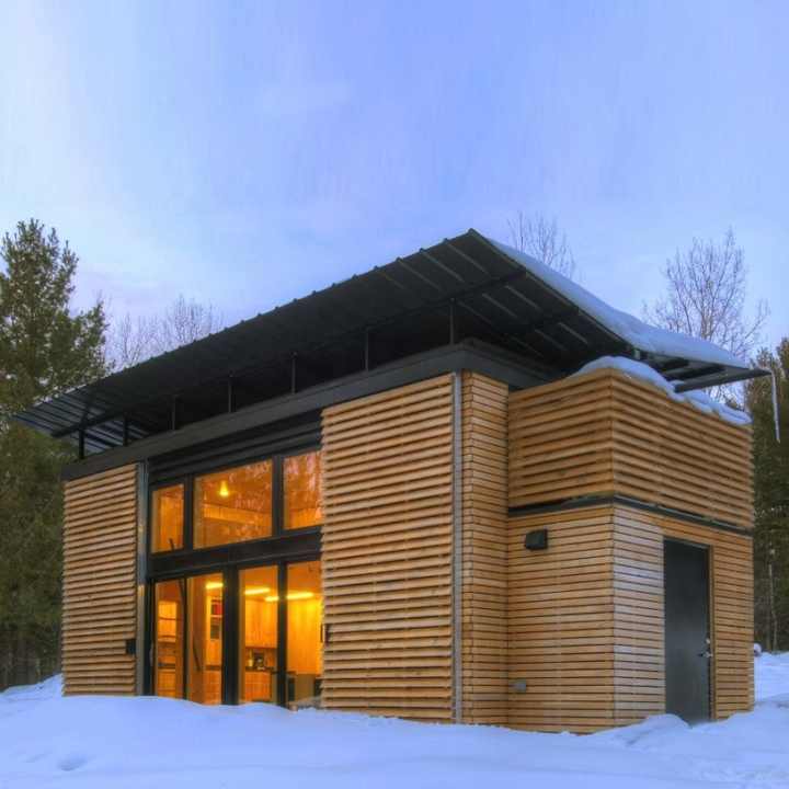 A modern, minimalist prefab house with a wooden slatted exterior and large glass windows, set in a snowy, forested landscape.