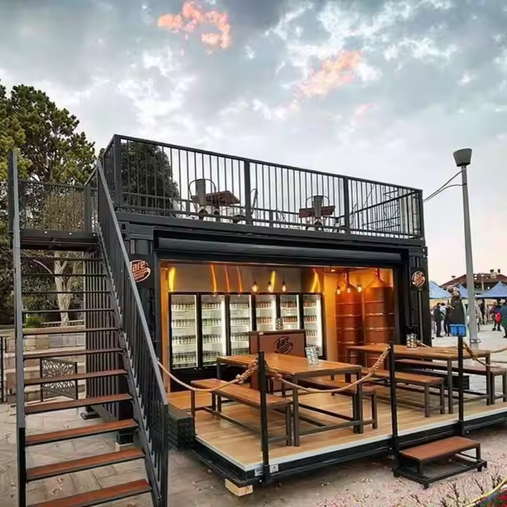 A minimalist, dark-toned prefab building with a flat roof and large windows, designed with clean lines and a small outdoor space, surrounded by sparse vegetation.